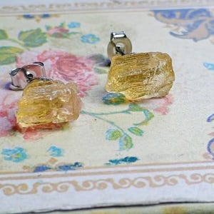 Puede incluir: Un par de pendientes de bot&oacute;n de citrino en bruto, de color amarillo dorado. Los pendientes tienen una textura natural y rugosa y est&aacute;n montados sobre postes de plata. Se presentan sobre una superficie con estampado floral.