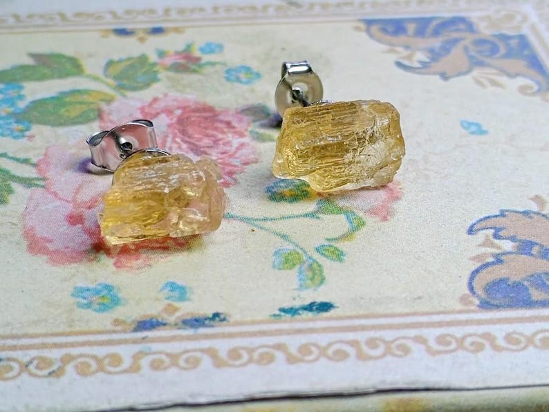 Puede incluir: Un par de pendientes de bot&oacute;n de citrino en bruto, de color amarillo dorado. Los pendientes tienen una textura natural y rugosa y est&aacute;n montados sobre postes de plata. Se presentan sobre una superficie con estampado floral.