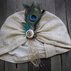 Può includere: Un turbante bianco con una piuma di pavone e un bottone decorativo. La piuma è blu, verde e nera. Il bottone è argentato e ha un motivo floreale.