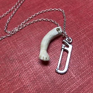 May include: A silver chain necklace with a pendant featuring a small, bone-colored arm and a silver saw charm. The necklace rests on a red textured surface.