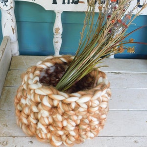 May include: A large, chunky knit basket made of white and brown yarn. The basket is filled with dried flowers and is sitting on a white wooden bench.
