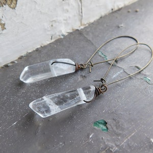 May include: A pair of earrings with clear quartz crystals and wire wrapped in copper. The earrings are hanging from a rustic background.