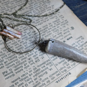 May include: A necklace featuring a long, pointed antler pendant and a crystal charm. The antler is light brown and the crystal is iridescent with pink and white tones. The necklace is made with a brown chain and a circular metal ring.