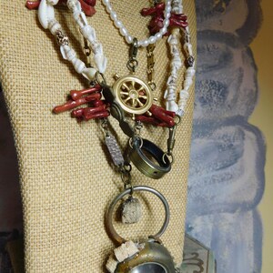 May include: A multi-strand necklace with white and red beads, a gold ship's wheel charm, a brass ring, and a round, rusty metal pendant with a clear glass window.