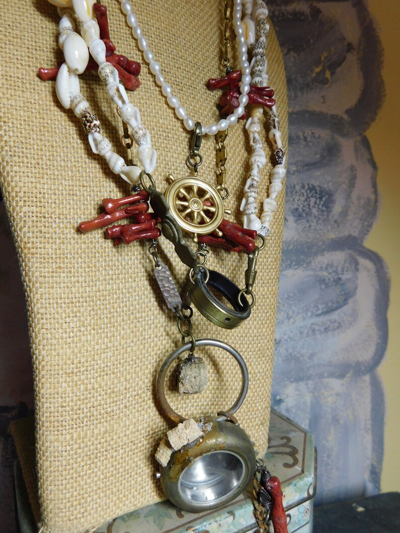 May include: A multi-strand necklace with white and red beads, a gold ship's wheel charm, a brass ring, and a round, rusty metal pendant with a clear glass window.