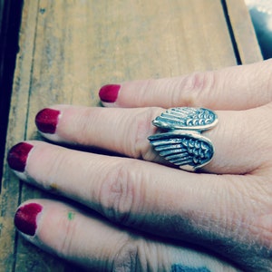 May include: A silver ring with a wing design. The ring is worn on a person's finger.
