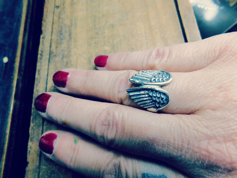 May include: A silver ring with a wing design. The ring is worn on a person's finger.