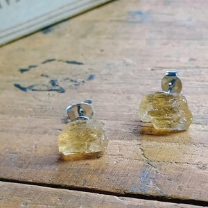 Puede incluir: Un par de pendientes de bot&oacute;n de citrino en bruto. Cada pendiente presenta un cristal de citrino amarillo dorado en bruto montado sobre un poste de plata. Los pendientes se muestran sobre una superficie de madera.