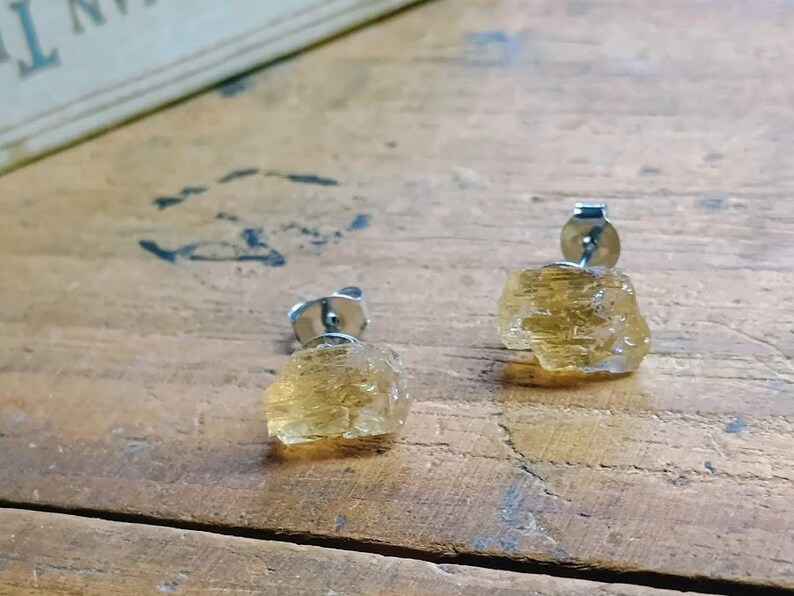 Puede incluir: Un par de pendientes de bot&oacute;n de citrino en bruto. Cada pendiente presenta un cristal de citrino amarillo dorado en bruto montado sobre un poste de plata. Los pendientes se muestran sobre una superficie de madera.