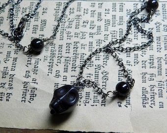 May include: A black chain necklace with a skull pendant and black beads. The necklace is lying on a page of an old book.