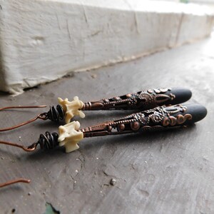 May include: A pair of long, black and copper earrings with a white bone-like charm at the top. The earrings have a detailed, intricate design.