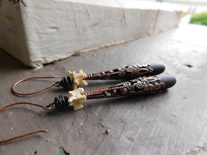 May include: A pair of long, black and copper earrings with a white bone-like charm at the top. The earrings have a detailed, intricate design.