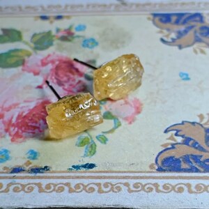 Puede incluir: Un par de pendientes de bot&oacute;n de piedra preciosa de color amarillo dorado en bruto. Los pendientes tienen forma cuadrada y tienen una superficie rugosa y texturizada. Est&aacute;n colocados sobre postes negros. Los pendientes se muestran sobre un fondo con estampado floral.