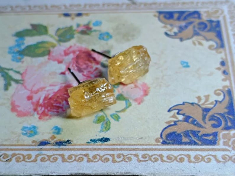 Puede incluir: Un par de pendientes de bot&oacute;n de piedra preciosa de color amarillo dorado en bruto. Los pendientes tienen forma cuadrada y tienen una superficie rugosa y texturizada. Est&aacute;n colocados sobre postes negros. Los pendientes se muestran sobre un fondo con estampado floral.