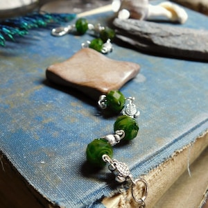 May include: A green and silver bracelet with a brown rectangular charm. The bracelet is made with green glass beads and silver spacers. The charm is made of wood and has a natural finish.