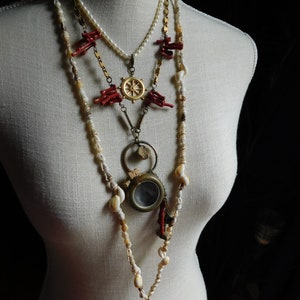 May include: A layered necklace with multiple strands of beads, including white pearls, red coral, and small white shells. The necklace features a gold compass charm and a small antique-looking magnifying glass.