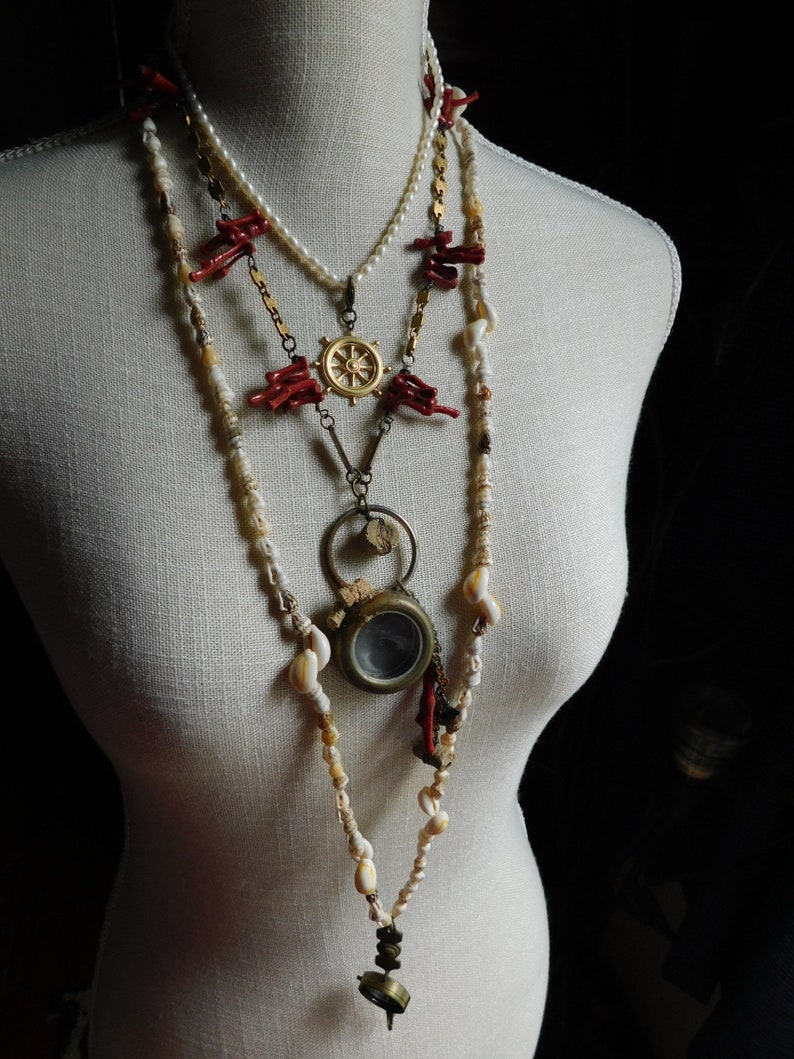 May include: A layered necklace with multiple strands of beads, including white pearls, red coral, and small white shells. The necklace features a gold compass charm and a small antique-looking magnifying glass.