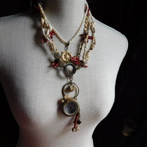 May include: A layered necklace with a variety of beads, including white pearls, red coral, and gold accents. The necklace features a pendant with a small, round, clear glass window.