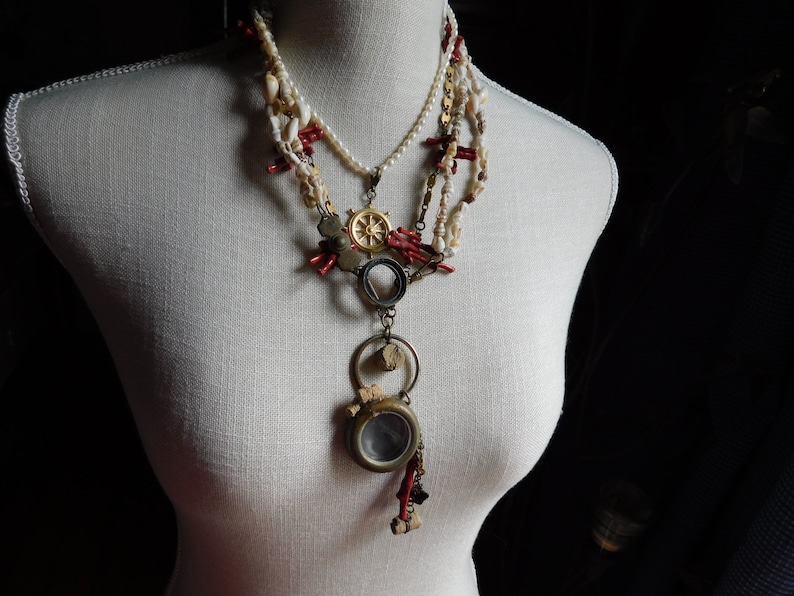 May include: A layered necklace with a variety of beads, including white pearls, red coral, and gold accents. The necklace features a pendant with a small, round, clear glass window.