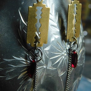 May include: A pair of earrings featuring a gold-toned razor blade charm with a red glass bead dangling from a chain.