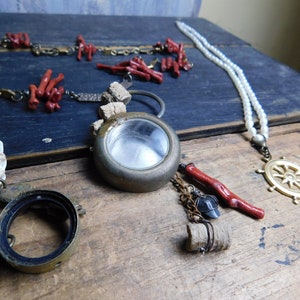 May include: Three necklaces with various charms and beads. One necklace features a round brass locket with a clear glass window, another has a white pearl strand with a gold ship's wheel pendant, and the third has red coral beads and a brass key.