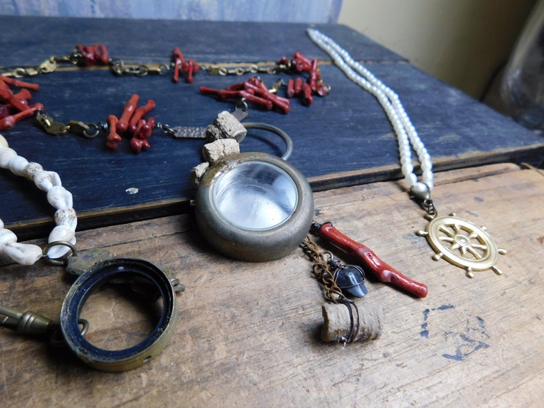 May include: Three necklaces with various charms and beads. One necklace features a round brass locket with a clear glass window, another has a white pearl strand with a gold ship's wheel pendant, and the third has red coral beads and a brass key.