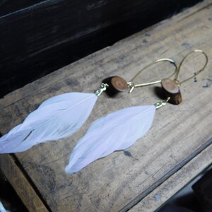 May include: A pair of earrings with a light pink feather charm and a wood accent. The earrings are made with gold-toned metal.