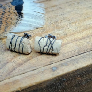 May include: A pair of handmade stud earrings. Each earring features a small, beige cork cylinder wrapped with dark wire. The earrings are set against a rustic wooden surface, with a feather in the background.