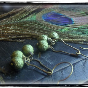 Puede incluir: Un par de pendientes con cuentas redondas verdes texturizadas y ganchos dorados. Los pendientes se muestran sobre una superficie oscura con una pluma de pavo real en el fondo.