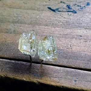 Puede incluir: Dos pendientes de bot&oacute;n de piedra preciosa amarillo claro, tallados en bruto. Los pendientes est&aacute;n montados sobre postes de metal oscuro, sobre una superficie de madera envejecida. Las piedras tienen un aspecto cristalino.