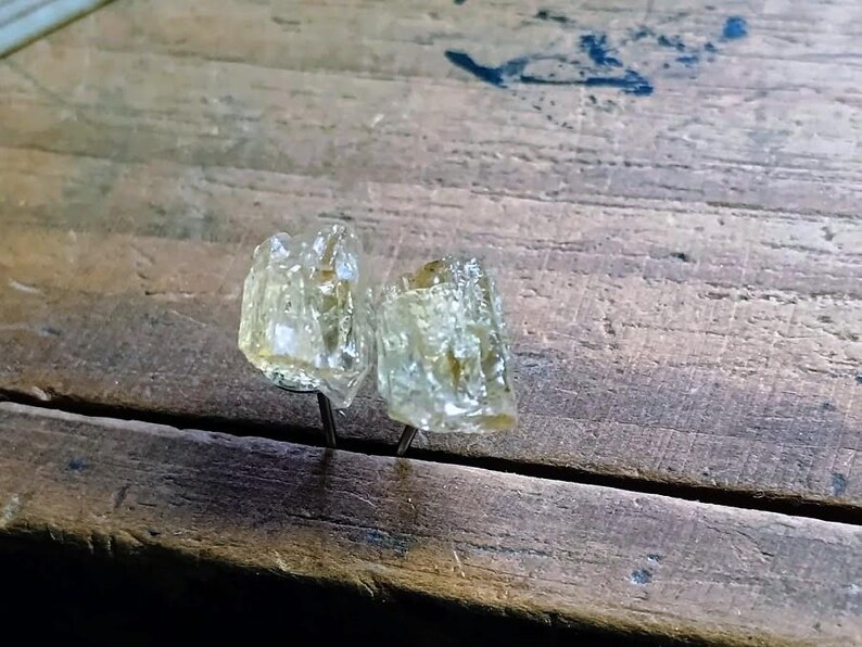 Puede incluir: Dos pendientes de bot&oacute;n de piedra preciosa amarillo claro, tallados en bruto. Los pendientes est&aacute;n montados sobre postes de metal oscuro, sobre una superficie de madera envejecida. Las piedras tienen un aspecto cristalino.