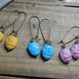 May include: Three pairs of dangle earrings with a skull design. The earrings are made of copper wire and have a small, oval-shaped bead with a white skull design. The beads are yellow, blue, and pink.