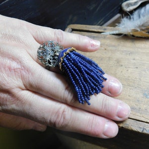 Op de afbeelding: Een zilveren ring met een filigraan ontwerp en een blauwe kwastbedel.