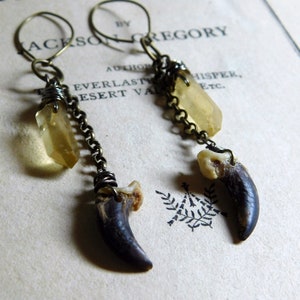 Puede incluir: Pendientes artesanales con un colgante de garra oscura, un cristal amarillo y una cadena. Los pendientes están hechos con metal color latón antiguo y tienen un diseño único inspirado en la naturaleza.