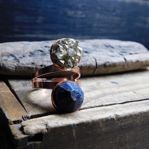 May include: A copper ring with two stones, one is a dark blue lapis lazuli and the other is a light brown pyrite. The ring is on a wooden surface.