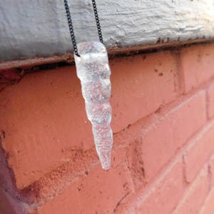 May include: A clear glass icicle pendant necklace with a black chain. The icicle is hanging from a brick wall.