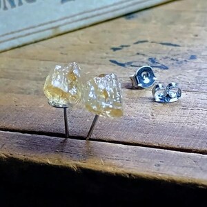 Puede incluir: Un par de pendientes de bot&oacute;n de citrino en bruto con postes y cierres de plata. Los pendientes tienen una textura rugosa y natural y un color amarillo dorado transl&uacute;cido. Los pendientes se exhiben sobre una superficie de madera.