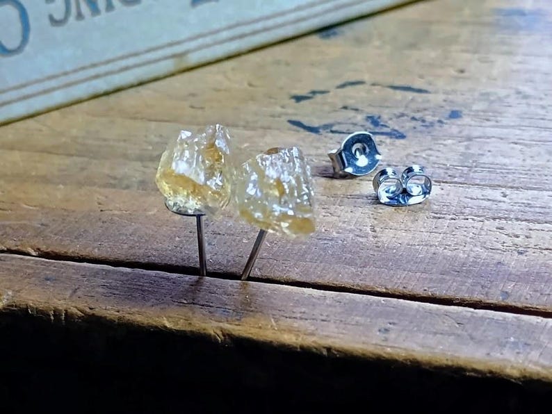 Puede incluir: Un par de pendientes de bot&oacute;n de citrino en bruto con postes y cierres de plata. Los pendientes tienen una textura rugosa y natural y un color amarillo dorado transl&uacute;cido. Los pendientes se exhiben sobre una superficie de madera.