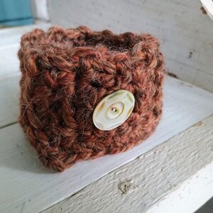 May include: A cylindrical crochet cup cozy made of thick, textured yarn in shades of rust, brown, and dark orange. The cozy features a single, round, cream-colored button with four holes attached to the front, securing the knitted fabric together.