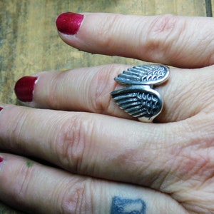 May include: A silver ring with a detailed wing design. The ring is worn on a finger with red nail polish.