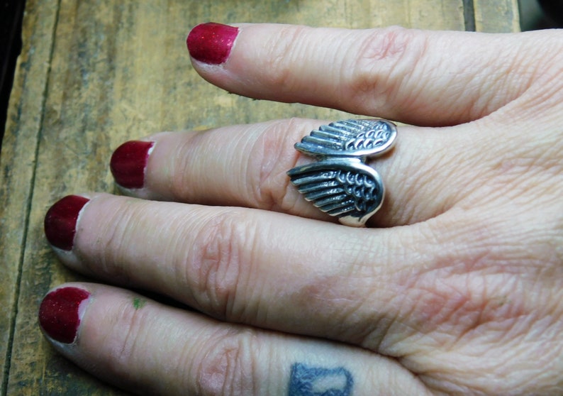 May include: A silver ring with a detailed wing design. The ring is worn on a finger with red nail polish.