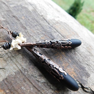 May include: A pair of earrings with black teardrop-shaped pendants and intricate copper filigree details. The earrings are attached to a copper wire with a white bone charm.
