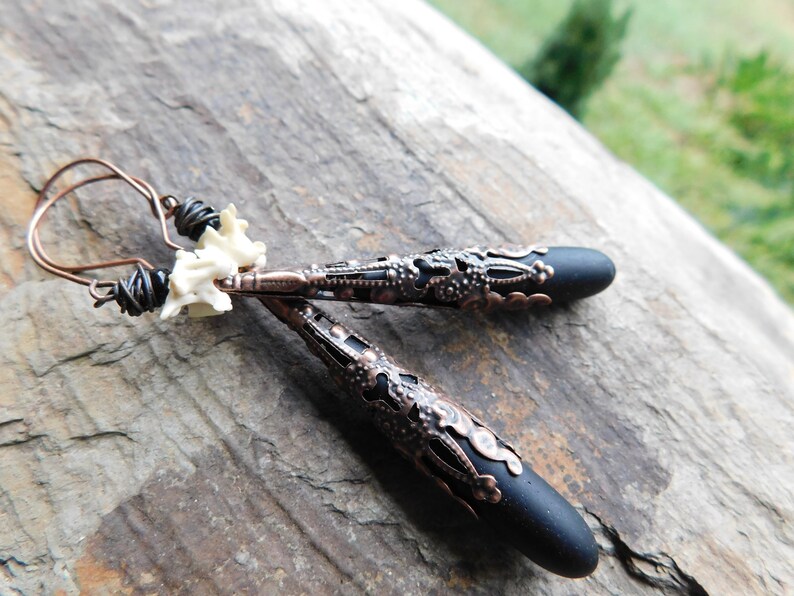 May include: A pair of earrings with black teardrop-shaped pendants and intricate copper filigree details. The earrings are attached to a copper wire with a white bone charm.