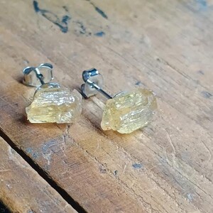 Puede incluir: Un par de pendientes de bot&oacute;n con cristales de citrino amarillo claro en bruto. Los pendientes tienen postes y cierres plateados. Los cristales tienen una textura natural rugosa, sobre una superficie de madera.