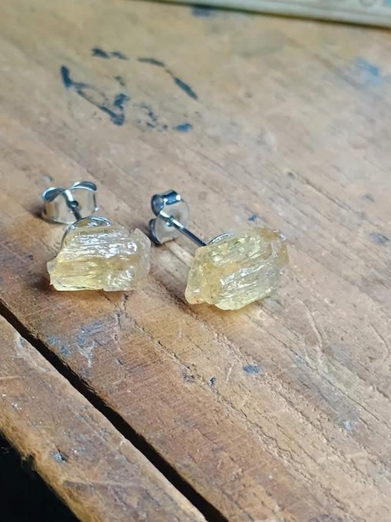 Puede incluir: Un par de pendientes de bot&oacute;n con cristales de citrino amarillo claro en bruto. Los pendientes tienen postes y cierres plateados. Los cristales tienen una textura natural rugosa, sobre una superficie de madera.