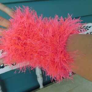 May include: A bright pink fuzzy wrist warmer made of yarn. The wrist warmer is worn on a person's arm.