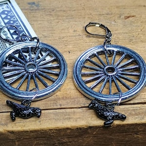 May include: A pair of silver-toned earrings featuring a wagon wheel design with black crow charms. The earrings are set against a wooden surface, showcasing their intricate details and craftsmanship. The earrings have a lever back closure.