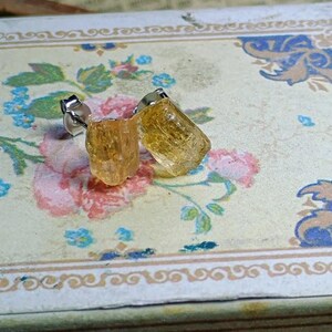 Puede incluir: Un par de pendientes de bot&oacute;n con piedras preciosas de color amarillo dorado en bruto. Los pendientes est&aacute;n engarzados en postes de plata y descansan sobre una superficie con estampado floral con detalles en rosa y azul.