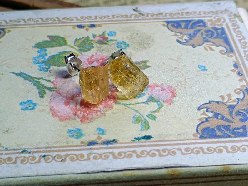 Puede incluir: Un par de pendientes de bot&oacute;n con piedras preciosas de color amarillo dorado en bruto. Los pendientes est&aacute;n engarzados en postes de plata y descansan sobre una superficie con estampado floral con detalles en rosa y azul.