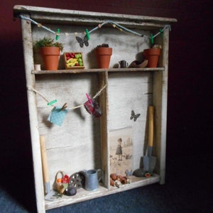 Puede incluir: Una estantería de madera blanca con dos estantes, decorada con plantas en miniatura, mariposas y herramientas de jardinería. El estante superior tiene una cuerda de cuentas azules y blancas con pinzas de ropa verdes. El estante inferior tiene una cuerda con un paño azul y un paño rojo colgando.